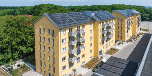 Flygfoto av ett modernt lägenhetskvarter med solpaneler på taken, omgivet av grönska.